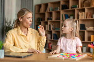 Caring mother teaching small preschool child daughter pronouncing words and letters correctly. Smart girl doing vocal exercises with professional teacher, sitting at desk